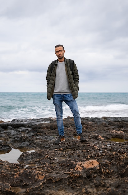 Homem posando no mar com atitude e uma mochila. Em um mar de pedras cercado por poças em um dia nublado. Ele está vestindo uma mochila, jeans, suéter e jaqueta verde. Suas mãos estão nos bolsos.