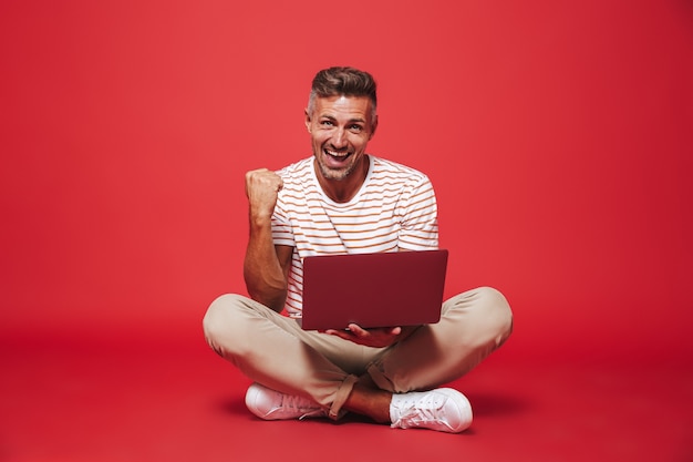 homem positivo de 30 anos em camiseta listrada sorrindo e usando laptop cinza, sentado no chão com as pernas cruzadas isoladas no vermelho