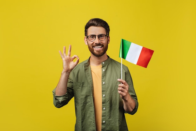 Foto homem positivo mostrando a bandeira da itália e gesticulando okey posando sobre fundo amarelo do estúdio e sorrindo para a câmera