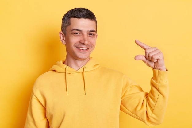Homem positivo otimista vestindo capuz estilo casual posando isolado sobre fundo amarelo mostrando um pouco de gesto polegada ou centímetro decepcionado com o tamanho mínimo
