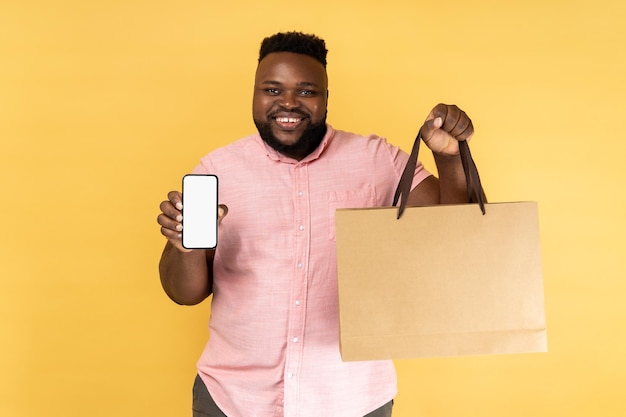 Homem positivo segurando sacola de compras mostrando celular com display vazio com área para propaganda