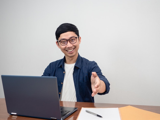 Homem positivo usa óculos sentados na mesa de trabalho apertando a mão para você