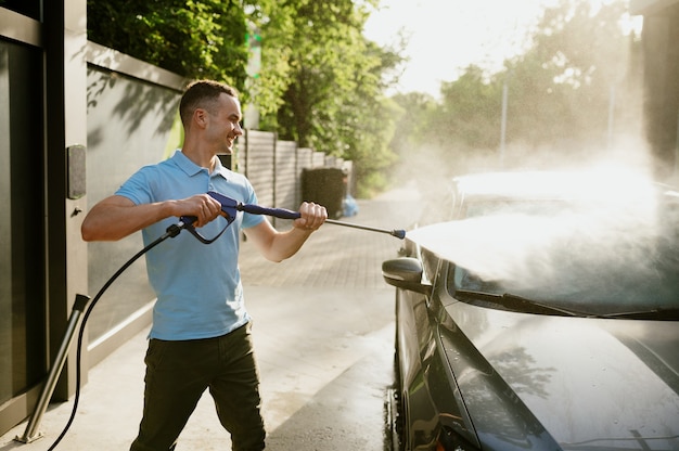 Homem possui pistola de água de alta pressão, estação de lavagem automática. indústria ou negócio de lavagem de carros. indivíduo do sexo masculino limpa seu veículo de sujeira ao ar livre