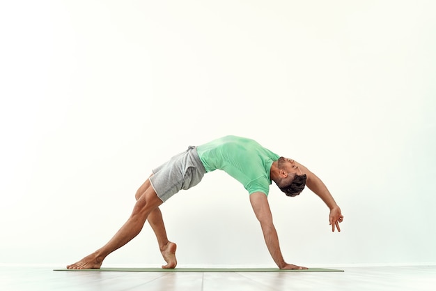 Homem praticando ioga em forma de pose selvagem camatkarasana contra uma parede branca