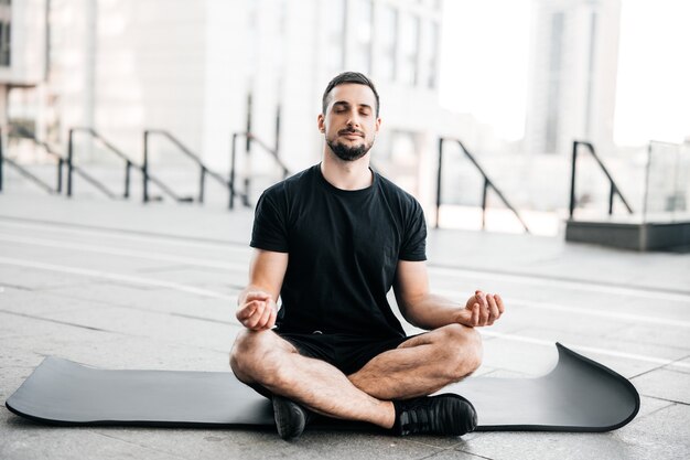 Homem praticando ioga na cidade grande. Homem desportivo meditando após o treino. Exercícios em casa. Jovem com barba por fazer, sentado no tapete de ioga preto com os olhos fechados ao ar livre e relaxante em pose de assento fácil.