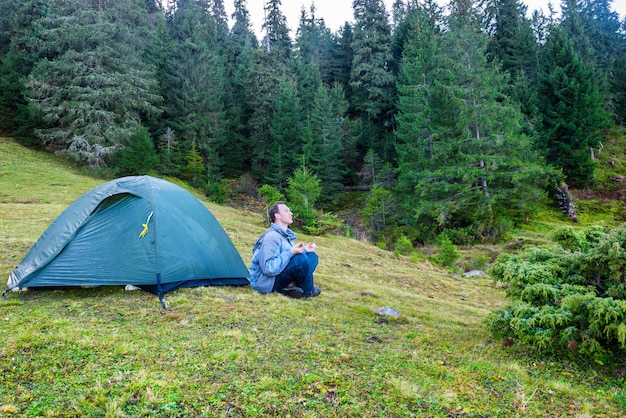 Homem praticando ioga perto da barraca de acampamento azul em uma floresta verde com pinheiros