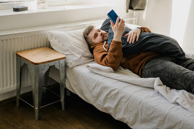 Foto homem preguiçoso usando o telefone enquanto está deitado na cama em casa