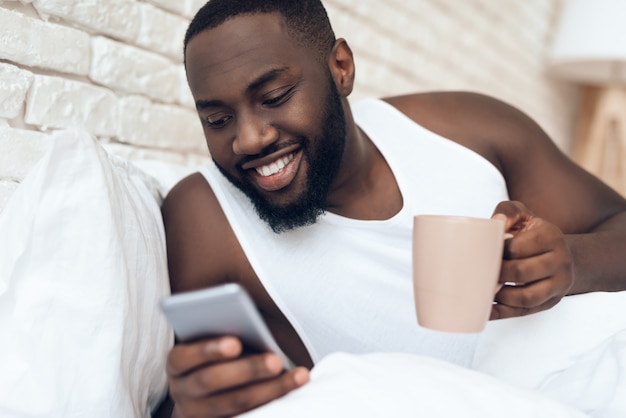 Homem preto, desperto bebe café na cama navegando.