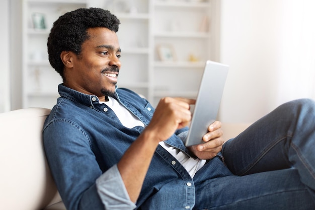 Homem preto sentado no sofá e relaxando com tablet digital