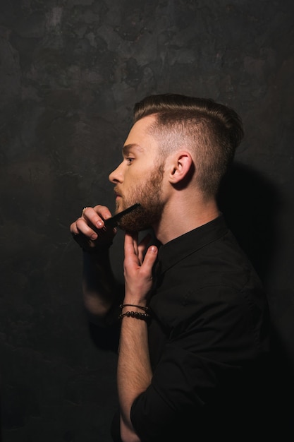 Homem puro hipster penteia a barba. Close de homem moderno e elegante em fundo escuro, retrato da juventude, conceito de beleza