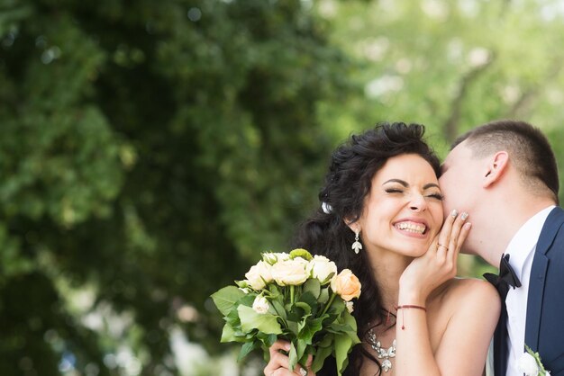 Foto homem recém-casado beija mulher feliz com buquê casal recém-casado sorri no verão ao ar livre