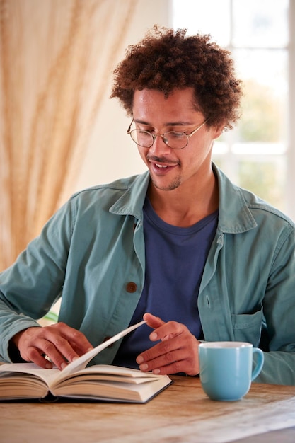 Homem relaxando em casa sentado à mesa lendo livro com bebida quente