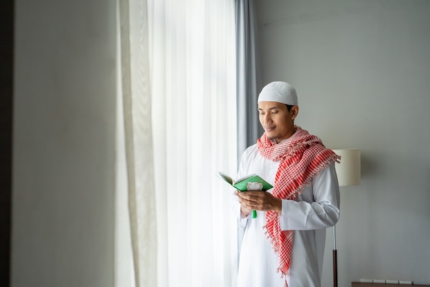 Homem religioso asiático lendo Alcorão ou Alcorão em pé ao lado da janela