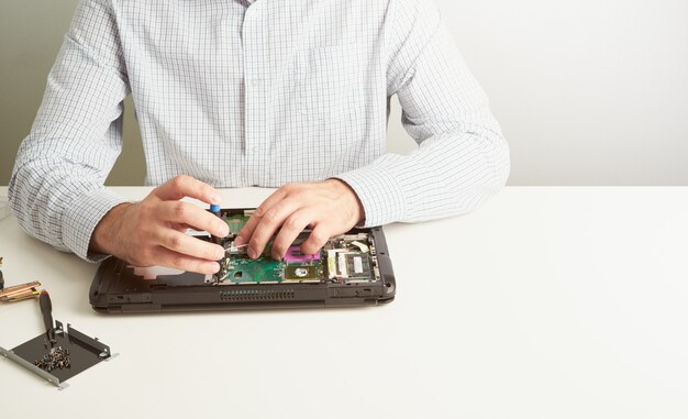 Homem repara o computador. Um engenheiro de serviço em camisa repara o laptop, na mesa branca contra a parede branca.