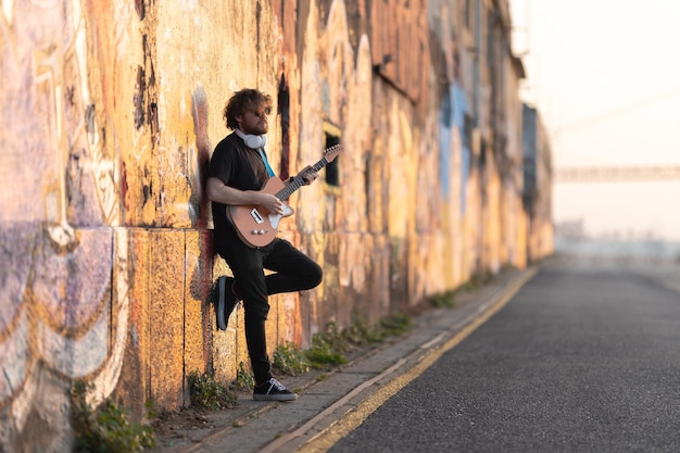 Foto homem romântico hipster usando óculos escuros em pé junto à parede tocando guitarra elétrica ao pôr do sol