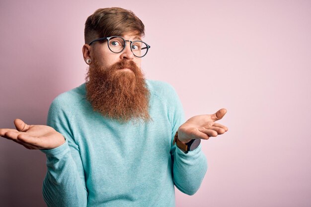 Foto homem roxo irlandês bonito com barba usando óculos sobre fundo rosa isolado expressão sem ideia e confusa com braços e mãos levantadas conceito de dúvida