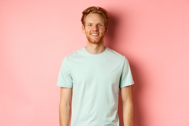 Homem ruiva feliz em t-shirt, olhando para a câmera e sorrindo, em pé sobre um fundo rosa.