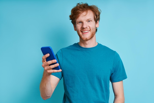 Homem ruivo alegre segurando o celular em pé contra o fundo azul