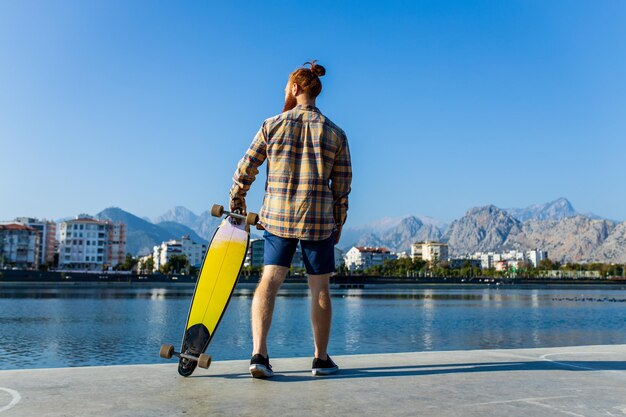Homem ruivo bonito com barba longa andando de skate perto do rio no dia ensolarado de verão