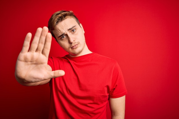 Homem ruivo bonito jovem vestindo camiseta casual sobre fundo vermelho isolado fazendo parar de cantar com a palma da mão Expressão de advertência com gesto negativo e sério no rosto