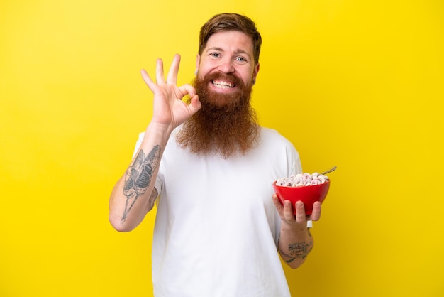 Homem ruivo com barba comendo uma tigela de cereais isolada em fundo amarelo, mostrando sinal de ok com os dedos
