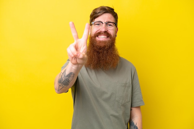 Homem ruivo com barba isolada em fundo amarelo sorrindo e mostrando sinal de vitória