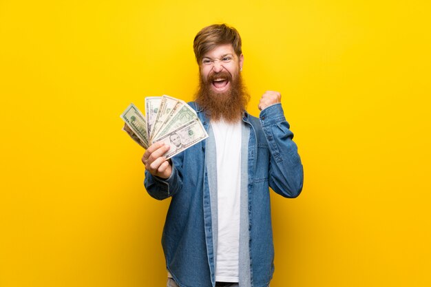 Homem ruivo com barba longa ao longo da parede amarela isolada, levando muito dinheiro