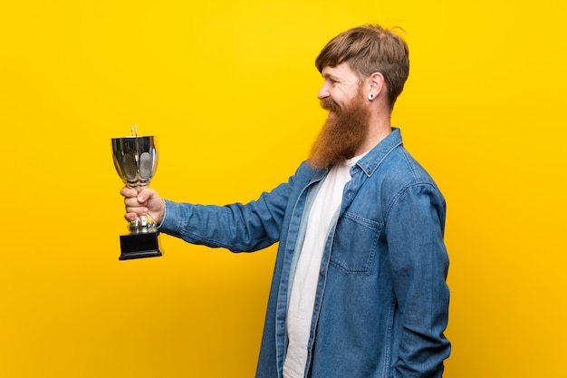 Homem ruivo com barba longa ao longo da parede amarela isolada, segurando um troféu