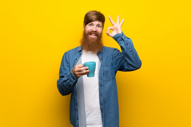Homem ruivo com barba longa ao longo da parede amarela, segurando a xícara de café quente