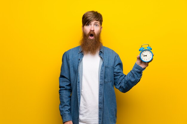 Homem ruivo com barba longa ao longo da parede amarela, segurando o despertador vintage