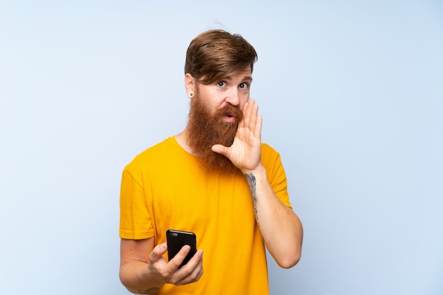 Homem ruivo com barba longa com um móvel sobre parede azul isolada, sussurrando algo