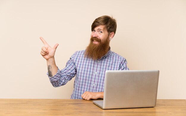 Homem ruivo com barba longa em uma mesa com um dedo apontando portátil para o lado