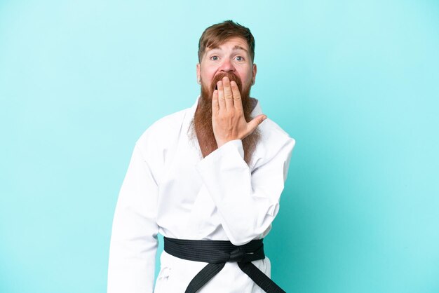 Homem ruivo com barba longa fazendo karatê isolado em fundo azul feliz e sorridente cobrindo a boca com a mão