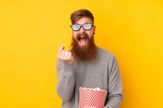 Homem ruivo com barba longa isolada parede amarela com óculos 3d e segurando um grande balde de pipocas