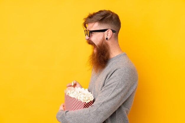 Homem ruivo com barba longa isolado parede amarela com óculos 3d e segurando um grande balde de pipocas enquanto olha de lado