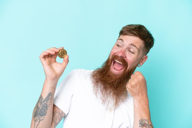 Homem ruivo com barba longa segurando um Bitcoin isolado no fundo azul comemorando uma vitória