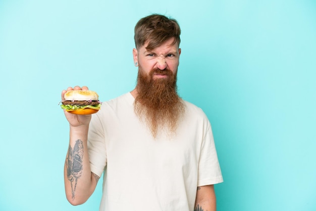 Homem ruivo com barba longa segurando um hambúrguer isolado no fundo azul com expressão triste