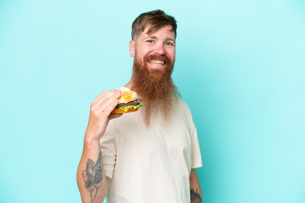 Homem ruivo com barba longa segurando um hambúrguer isolado no fundo azul sorrindo muito