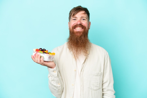 Homem ruivo com barba longa segurando uma tigela de frutas isolada no fundo azul sorrindo muito