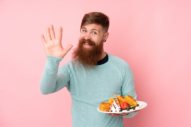 Homem ruivo com barba longa segurando waffles sobre parede rosa isolada, saudando com a mão com expressão feliz