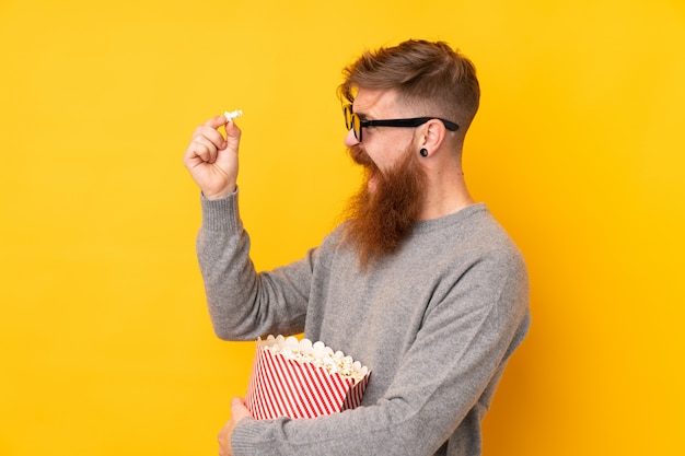 Homem ruivo com barba longa sobre amarelo isolado com óculos 3d e segurando um grande balde de pipocas