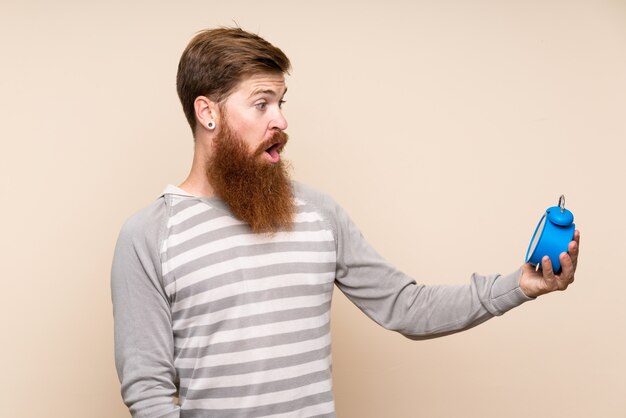 Homem ruivo com barba longa, sobre fundo isolado, segurando o despertador vintage