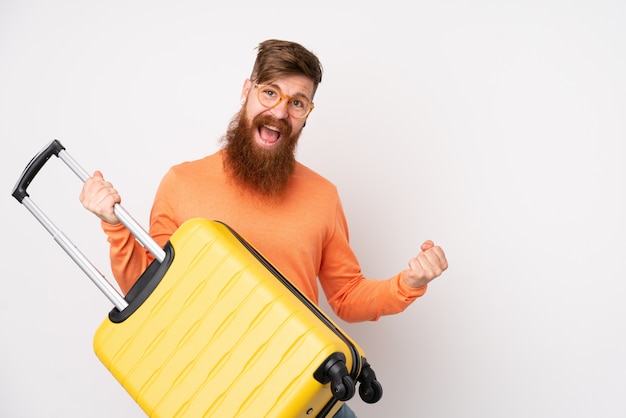 Homem ruivo com barba longa sobre parede branca isolada em férias segurando uma mala de viagem como uma guitarra