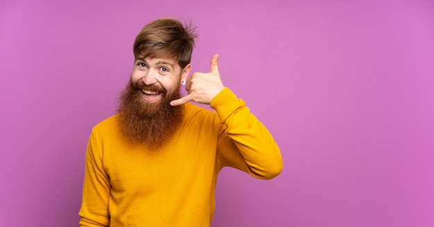 Homem ruivo com barba longa sobre parede roxa, fazendo gesto de telefone