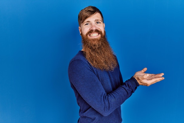 Homem ruivo com barba longa vestindo suéter azul casual sobre fundo azul apontando de lado com as palmas das mãos abertas mostrando espaço de cópia, apresentando anúncio sorrindo animado feliz