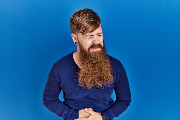 Homem ruivo com barba longa vestindo suéter azul casual sobre fundo azul com a mão no estômago porque indigestão, doença dolorosa se sentindo mal. conceito de dor.