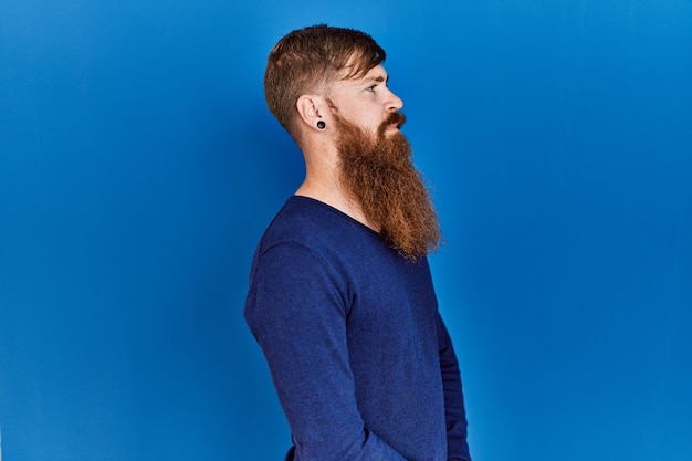 Homem ruivo com barba longa, vestindo suéter azul casual sobre fundo azul, olhando para o lado, relaxe a pose de perfil com rosto natural e sorriso confiante.