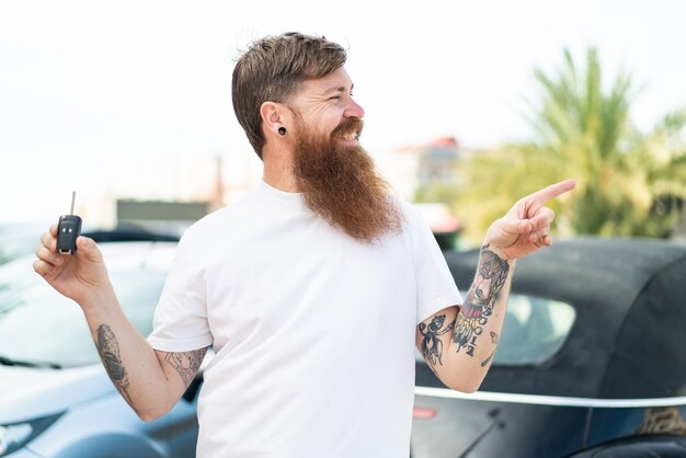 Homem ruivo com barba segurando as chaves do carro ao ar livre apontando para o lado para apresentar um produto