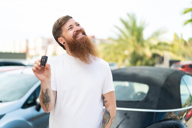 Homem ruivo com barba segurando as chaves do carro ao ar livre olhando para cima enquanto sorri