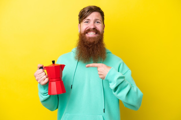 Homem ruivo com barba segurando o bule de café isolado no fundo amarelo e apontando-o